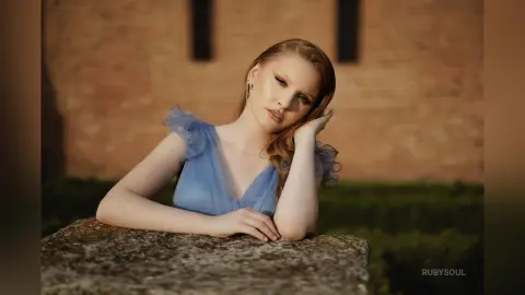 RubySoul poses leaning on a stone ledge wearing a light blue ruffled dress with her hand touching her hair.