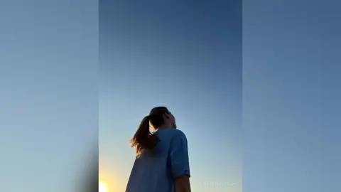 Dreamy silhouette of a woman in a blue shirt looking up at a clear blue sky during golden hour.