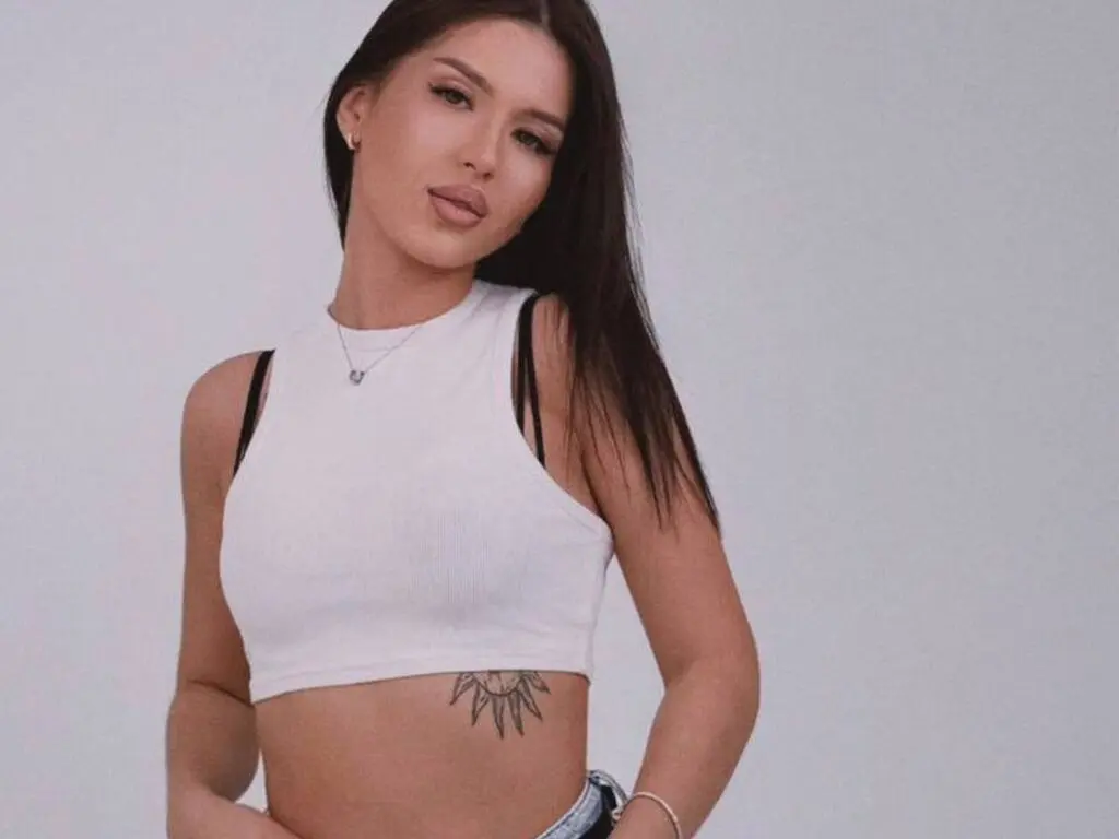 JadaJosey poses in a white crop top, showing her long dark hair and a tattoo on her stomach against a plain grey background.
