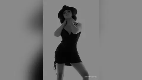 Moody black and white portrait of a woman in a chic black dress and wide-brimmed hat against a plain background.