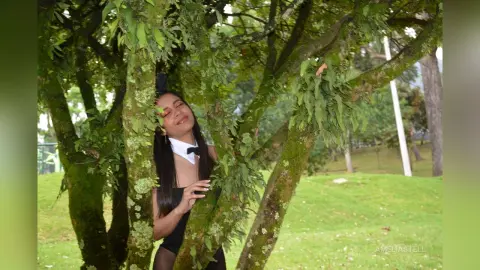 AmeliaStell poses behind a mossy tree trunk in a white collared shirt and black bowtie, looking up with eyes closed.