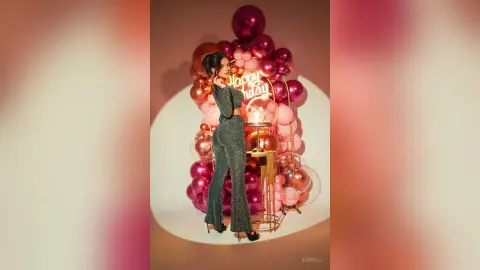 Glamorous woman in a sparkling silver jumpsuit posing by a pink balloon arch for a festive birthday celebration.