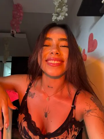 Smiling young woman in a black lace camisole with spider tattoos in a warm, romantic bedroom setting.