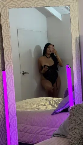 Young woman in a black sparkly bodysuit taking a selfie in a bedroom mirror with pink LED lighting.