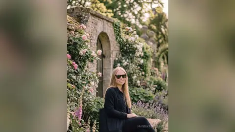Smiling blonde woman in a black outfit sitting in a lush blooming rose garden near a stone archway.
