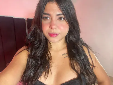 Smiling young woman with long wavy dark hair wearing a black tank top in a soft pink lit room.
