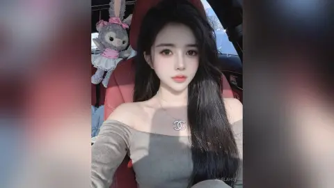 AgelaHui with long black hair wearing a grey off-the-shoulder top and a silver pendant, sitting in a red car seat next to a stuffed bunny.