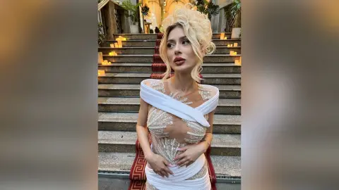 DaisyLin poses on a stone staircase wearing a white, sheer, wrap-style top that shows her cleavage and midriff.