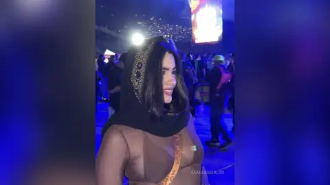 Smiling woman in a black embellished hijab and sheer brown top at a vibrant nighttime outdoor event.