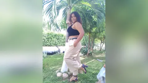 Young woman in a black crop top and beige trousers posing in a sunny tropical garden.