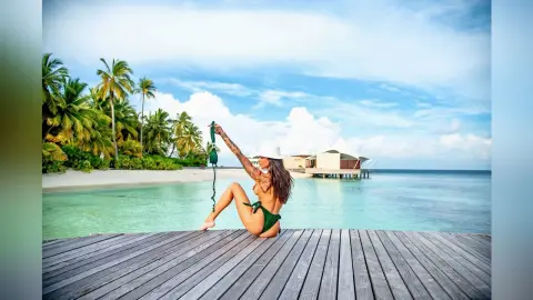Tattooed woman in a green bikini relaxing on a wooden pier overlooking a tropical turquoise ocean beach.