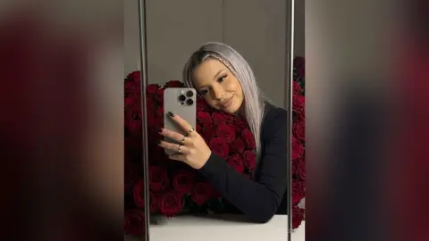 Kalira poses for a mirror selfie, wearing a black long-sleeve top with her long, silver-grey hair styled down against a background of red roses.