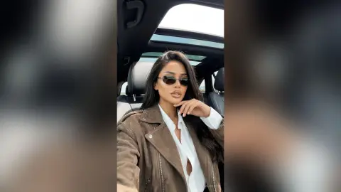 Stylish woman in sunglasses and a brown suede jacket sitting in a car with a sunroof on a bright day.