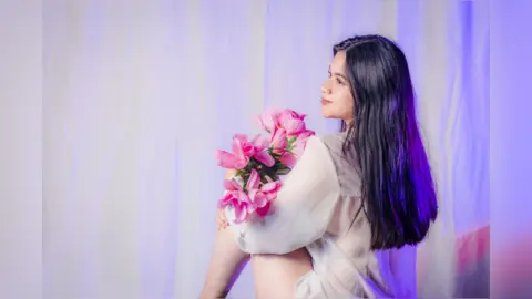 Young woman in a white blouse holding pink flowers against a soft purple backdrop in a dreamy mood.