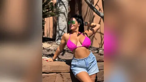 MissyJolie poses outdoors in a bright pink bikini top and denim shorts, smiling with her hand behind her head.