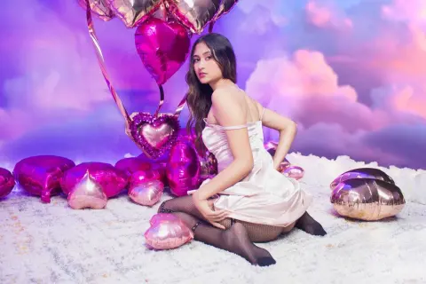 Dreamy portrait of a woman in a pink satin dress sitting among heart balloons in a whimsical cloud setting.
