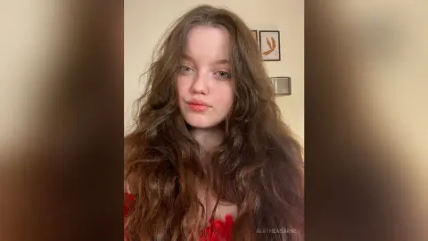 Young woman with long wavy brown hair wearing a red top in a bright, minimalist indoor setting.