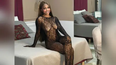 LiaTyler smiles while sitting on a bed wearing a long-sleeved black lace bodysuit that shows her breasts and skin through the mesh.