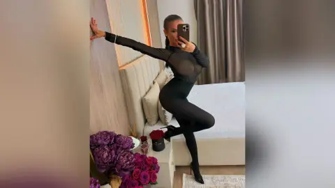 Stylish woman in a black long-sleeve bodysuit posing in a modern bedroom with purple flowers.