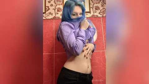 DeidreSarin poses in a purple button-down shirt and black pants, showing her midriff and navel piercing against a red tiled wall.