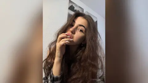 Young woman with long wavy hair in a black plaid shirt posing thoughtfully in a bright indoor setting.