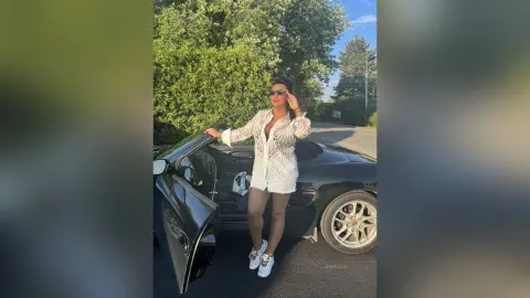 MissyJolie stands next to a black convertible car wearing a white lace shirt and white shorts.