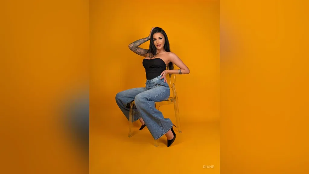 Tattooed woman in black tube top and blue jeans sitting against a vibrant yellow background.