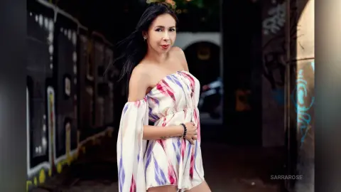 Young woman in a white and purple floral off-the-shoulder dress posing in a dark urban alleyway.