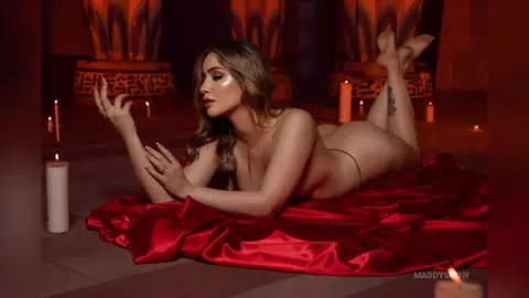 Glamorous blonde woman in gold lingerie lounging on red satin amidst glowing candles in a moody, romantic setting.