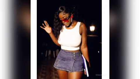 Stylish Black woman in a white tank top and denim mini skirt posing outdoors at night with red sunglasses.