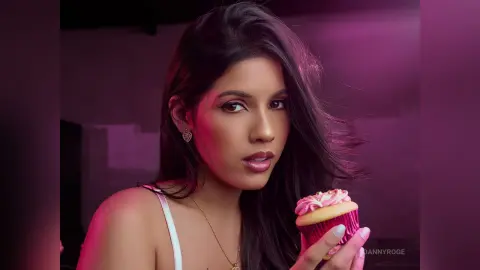 Young woman in white tank top holding a pink cupcake against a moody purple background.