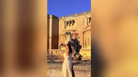 Woman in white dress standing before a classical stone fountain sculpture at golden hour.