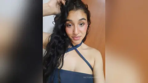 Young woman with long wavy hair wearing a blue halter neck top in a soft, natural indoor setting.