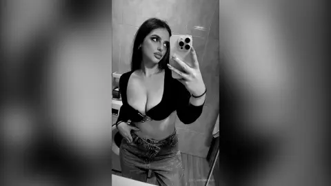 Young woman in a black crop top and denim jeans taking a moody black and white mirror selfie in a bathroom.