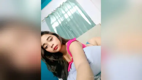 Young woman in a pink tank top relaxing in a bright bedroom with teal curtains.