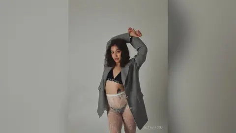 Young woman in a grey blazer and lace lingerie posing confidently against a minimalist grey background.