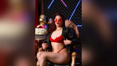 Glamorous woman in red lingerie and black leather jacket wearing red sunglasses in a neon-lit studio setting.