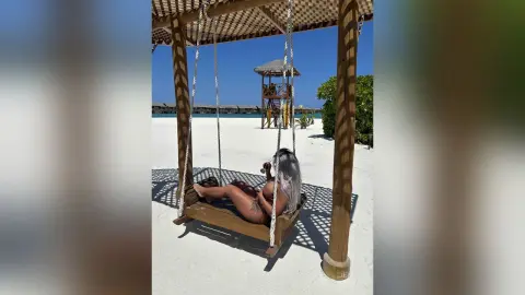 Blonde woman in a bikini relaxing on a wooden swing at a sunny tropical beach resort.