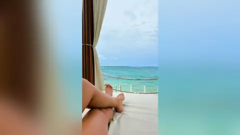 JulianaNavarro relaxing on a white bed or lounge chair outdoors by the turquoise ocean, showing her bare legs and feet.