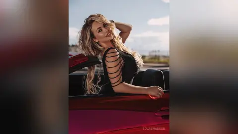 Glamorous blonde woman in a black cutout dress leaning against a red convertible car under bright sunlight.