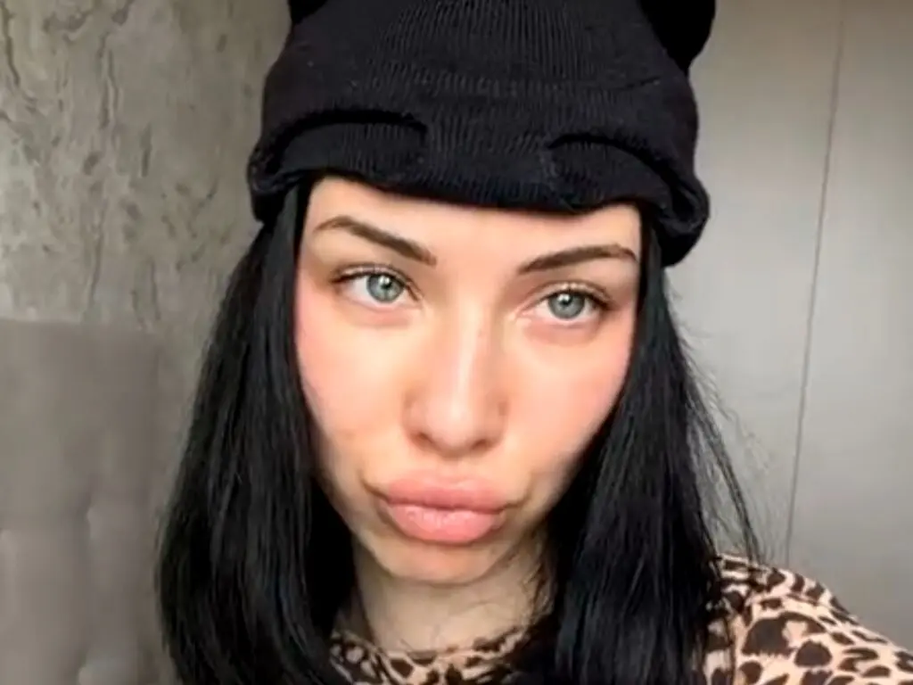 Young woman with long black hair and blue eyes wearing a black beanie and leopard print top in a soft indoor setting.