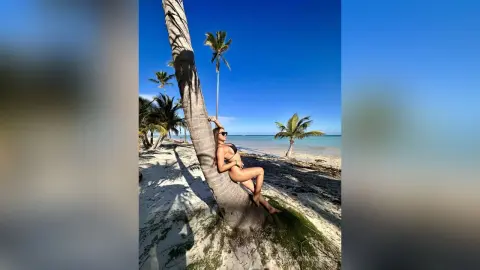 Smiling woman in a black bikini relaxing against a palm tree on a sunny tropical beach.