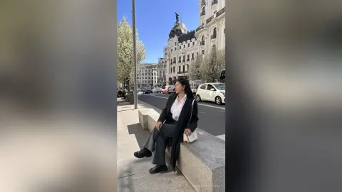 NashiroSakurai sits on a stone ledge wearing a long black coat, white top, and grey trousers in a bright outdoor city setting.