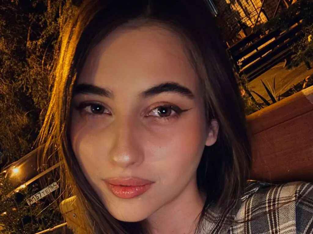 Young woman with winged eyeliner and plaid shirt smiling in warm nighttime outdoor lighting.