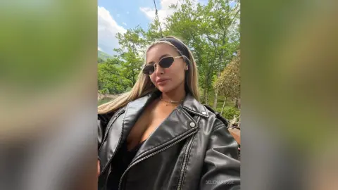 SelenaRusse wears sunglasses and a black leather jacket while posing outdoors against a backdrop of green trees.