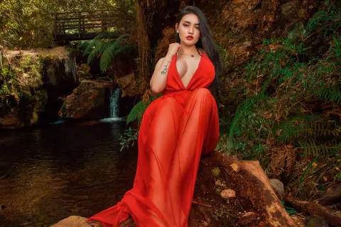 Elegant woman in a flowing red gown sitting by a forest waterfall in a dreamy, romantic woodland setting.