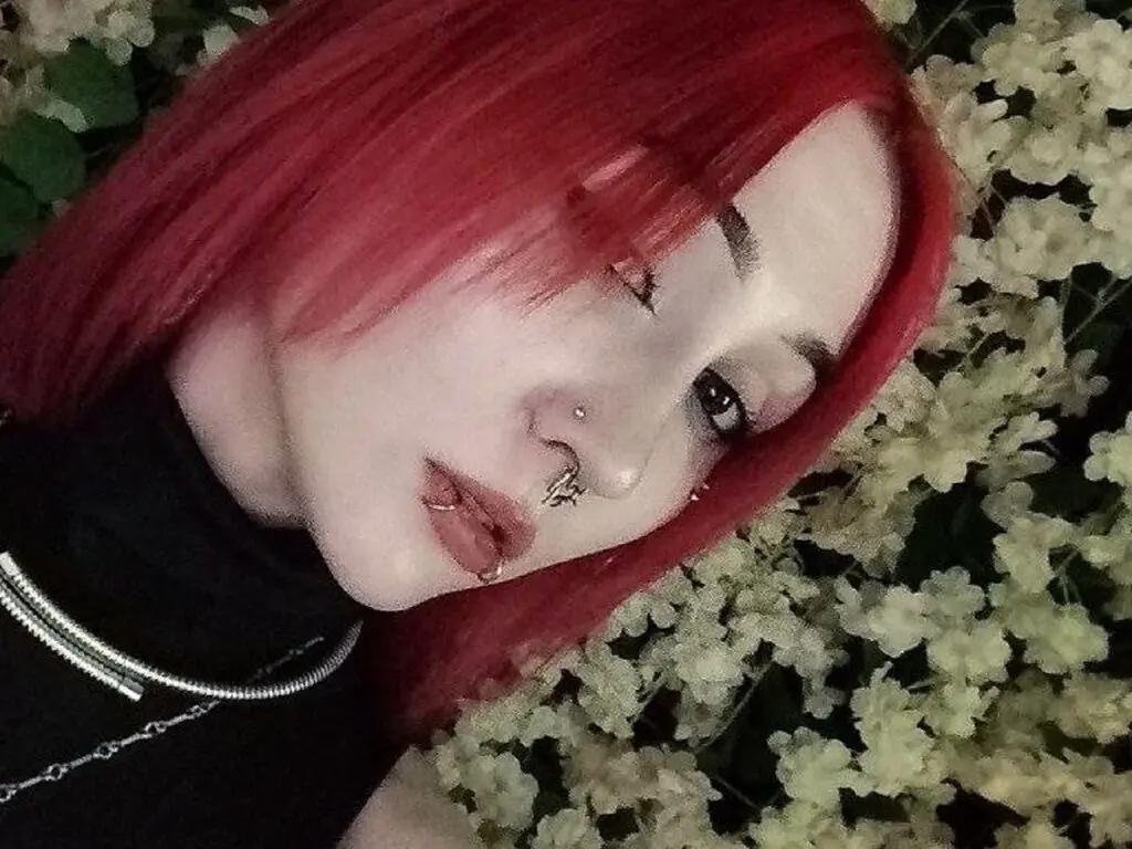 Close-up portrait of a woman with vibrant red hair and nose ring posing amidst white flowers in a moody setting.