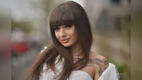 Smiling brunette woman with bangs in a white off-the-shoulder top posing outdoors in soft natural light.