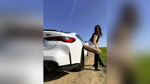 DyandraEve poses outdoors next to a white car, wearing a black top, white shorts, and black thigh-high boots.