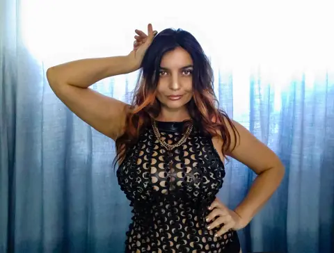 DominaPaulina poses in a black cutout dress with her hand in her hair against blue curtains.